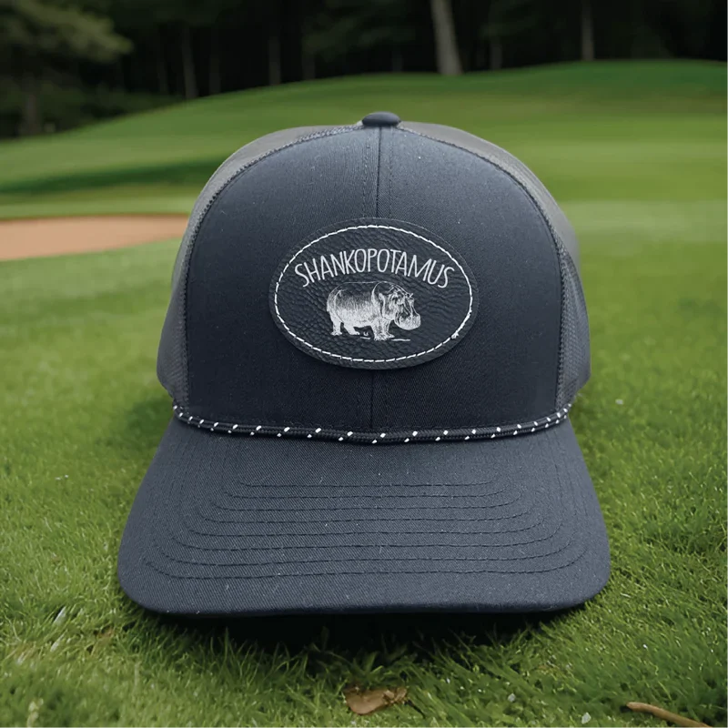 Dark gray trucker cap with embroidered oval patch reading "Shankopotamus" and hippo image, displayed on grass with baseball field background.