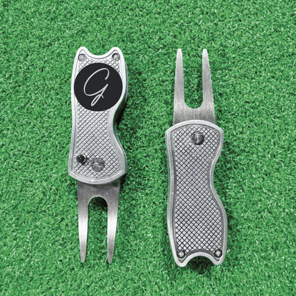 Two silver golf divot tools with textured handles on green turf, left tool featuring a black circle with sample monogram initial G