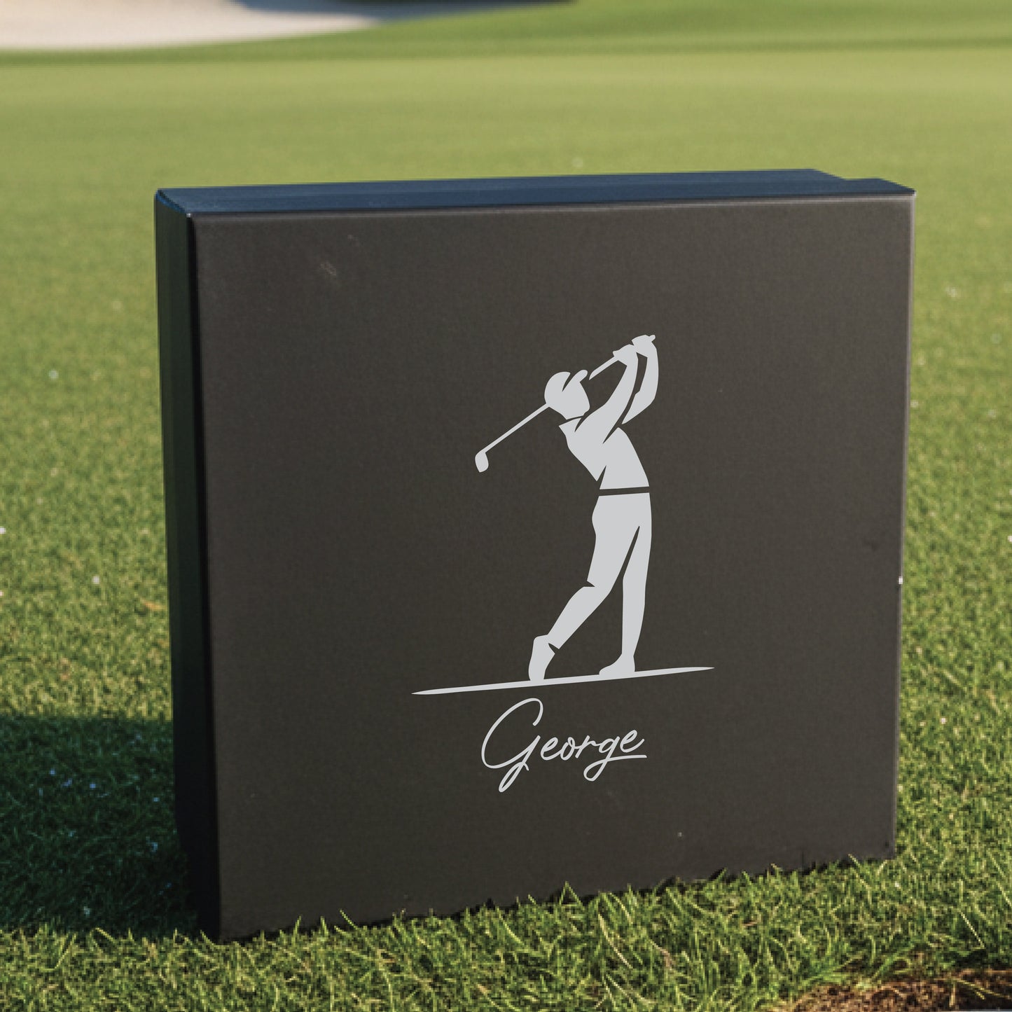 Black gift box with engraved golfer silhouette in mid-swing and sample name "George" on grass background
