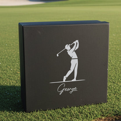 Black gift box with engraved golfer silhouette in mid-swing and sample name "George" on grass background