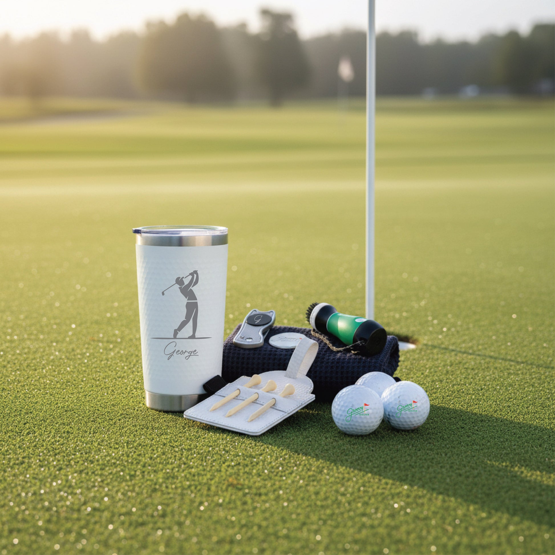 White insulated tumbler with sample personalization "George" and golfer silhouette, displayed on putting green with golf balls, tees, rangefinder, and black golf towel.