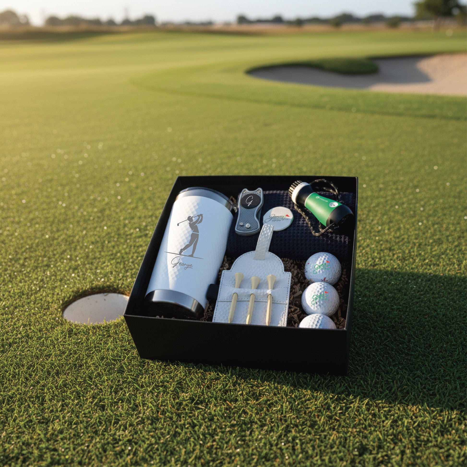 Black gift box on golf course containing engraved flask with golfer design, divot tool, golf balls, tees, and rangefinder with sample personalization.