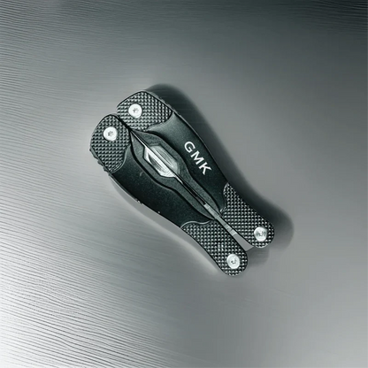 Black multi-tool with engraved "GMK" initials, textured grip handles, and multiple folded tools visible, displayed on gray brushed surface.