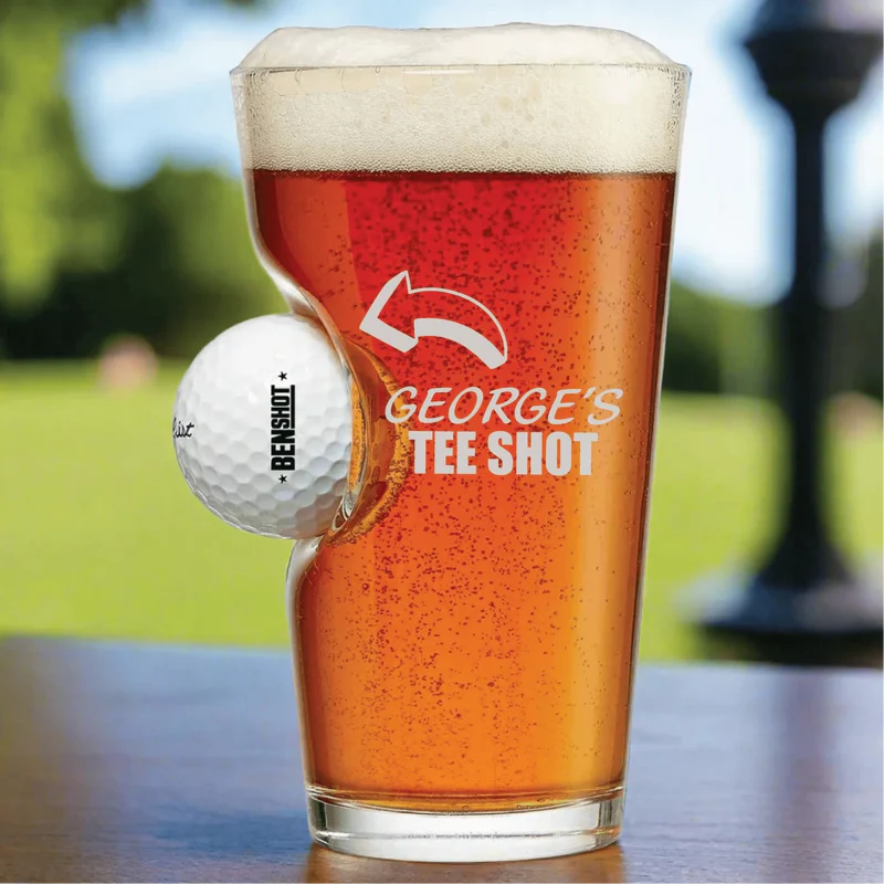 Engraved pint glass with sample text "George's Tee Shot" and golf ball design, filled with beer on a golf course background