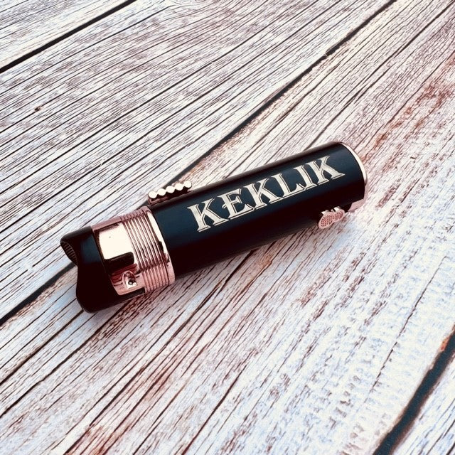 Black engraved flask with sample name "KEKLIK" and copper band detail on whitewashed wood surface.
