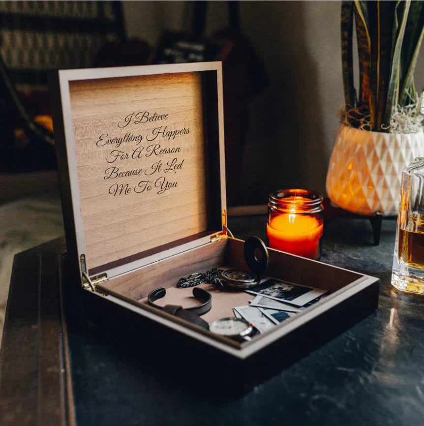 Open wooden gift box with engraved lid displaying sample text, containing cufflinks and accessories, displayed on dark surface with candle and glass.