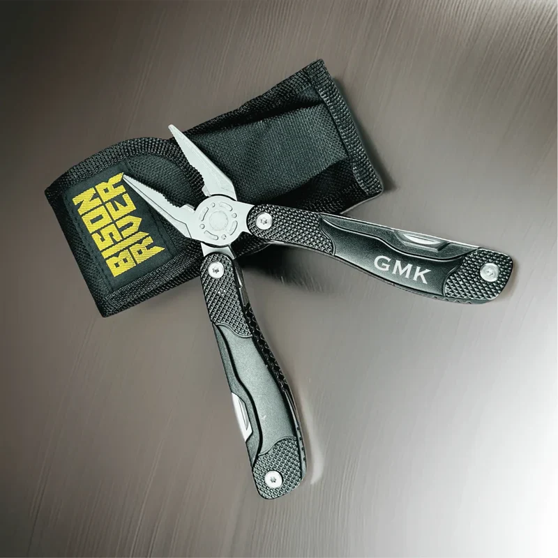 Engraved multi-tool knife with sample initials "GMK" on blade, black textured handles, and black nylon carrying pouch with yellow text sample.