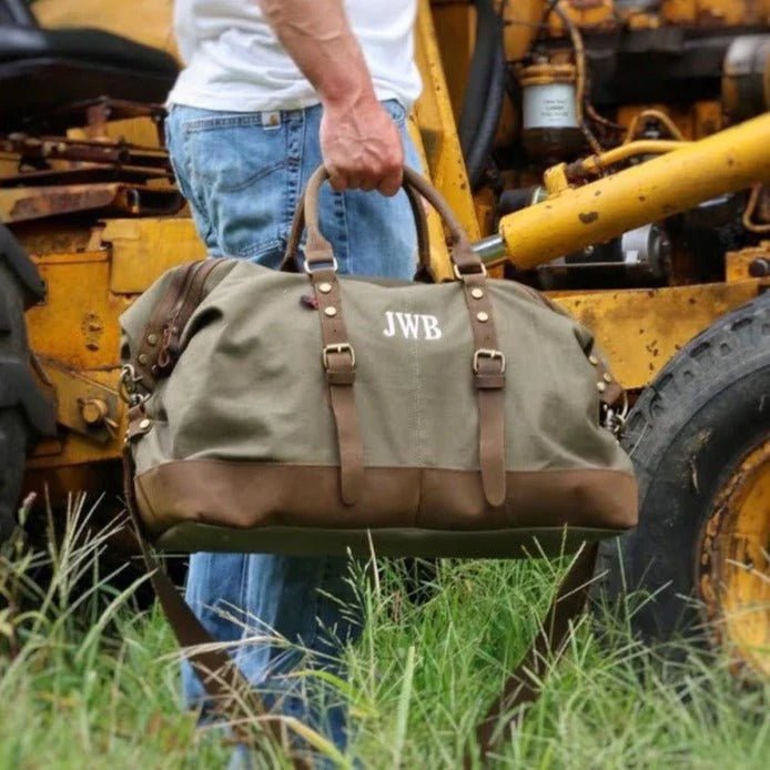 Person holding a green duffel bag with 'JWB' initials in front of a yellow piece of machinery.