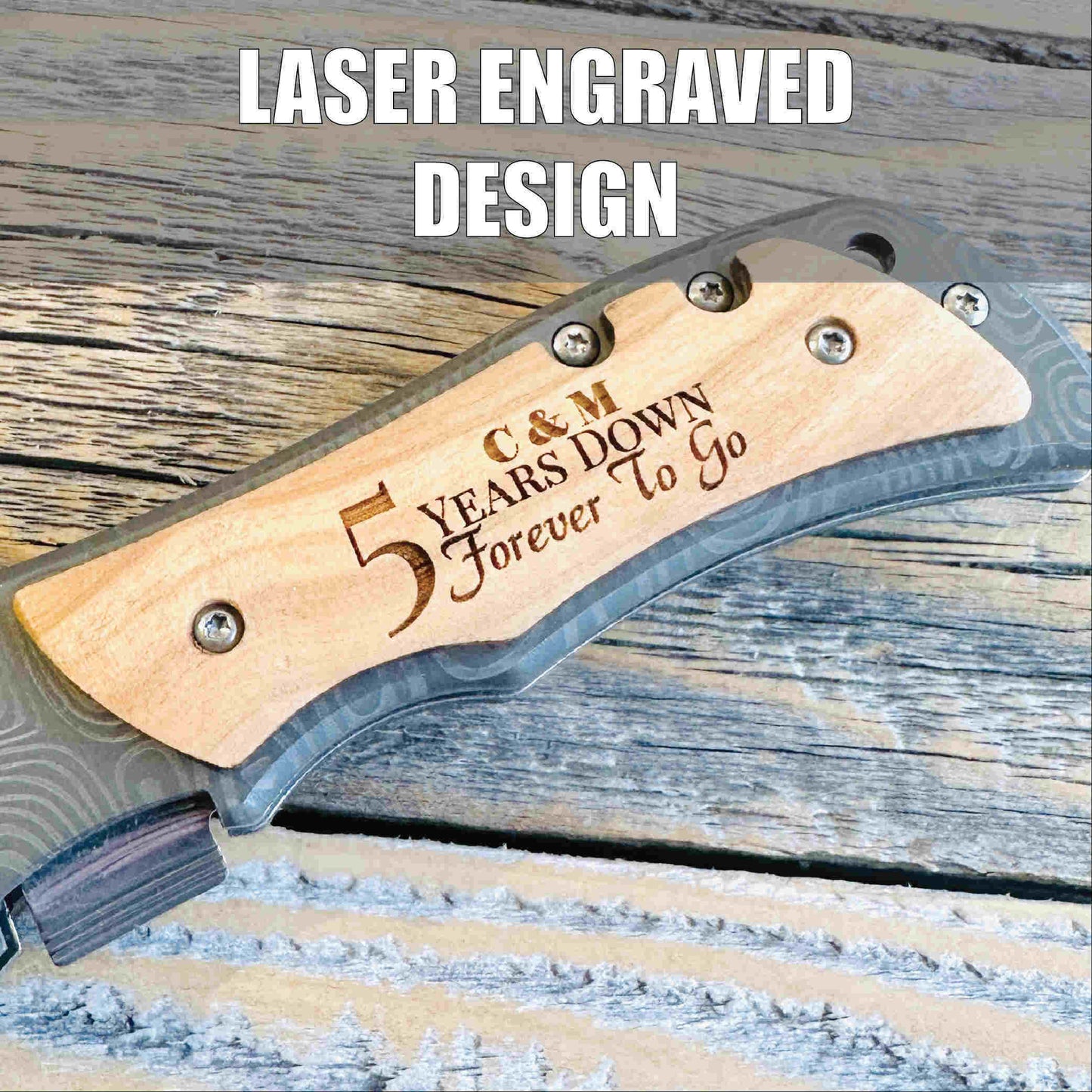 Folding knife with natural wood handle engraved with sample text "C & M 5 Years Down Forever To Go" on weathered wood surface.