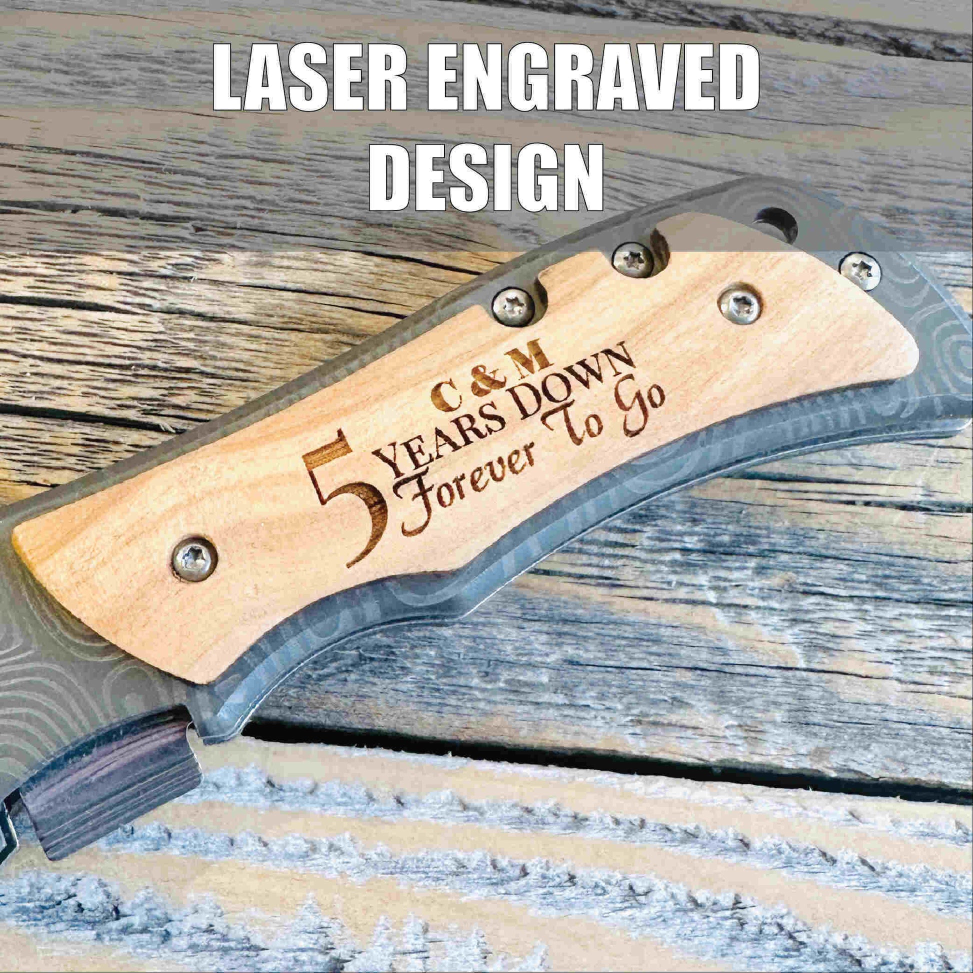 Folding knife with natural wood handle engraved with sample text "C & M 5 Years Down Forever To Go" on weathered wood surface.