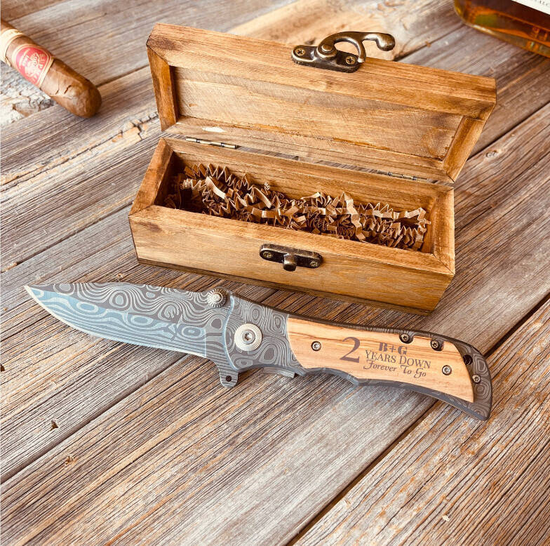Engraved folding knife with wood handle displaying "2 Years Down Forever To Go" sample text, displayed with open wooden gift box and cigar.