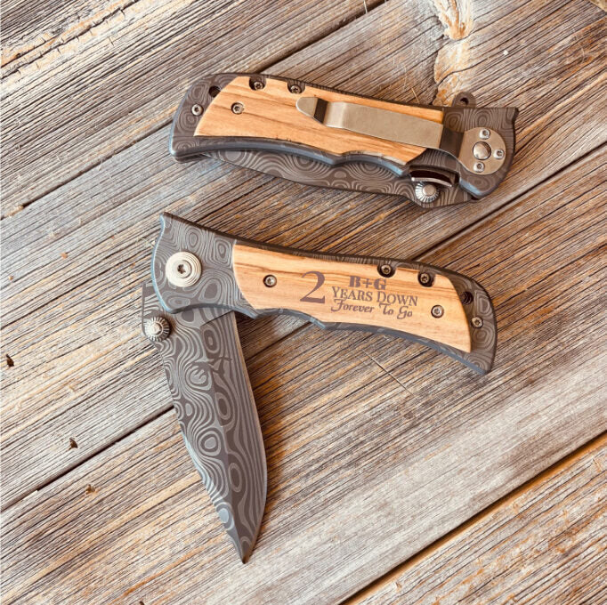 Two folding pocket knives with wood handles and patterned metal scales, bottom knife engraved with sample text "2 Years Down Forever to Go