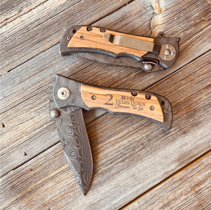 Two folding pocket knives with wood handles and patterned metal scales, bottom knife engraved with sample text "2 Years Down Forever to Go