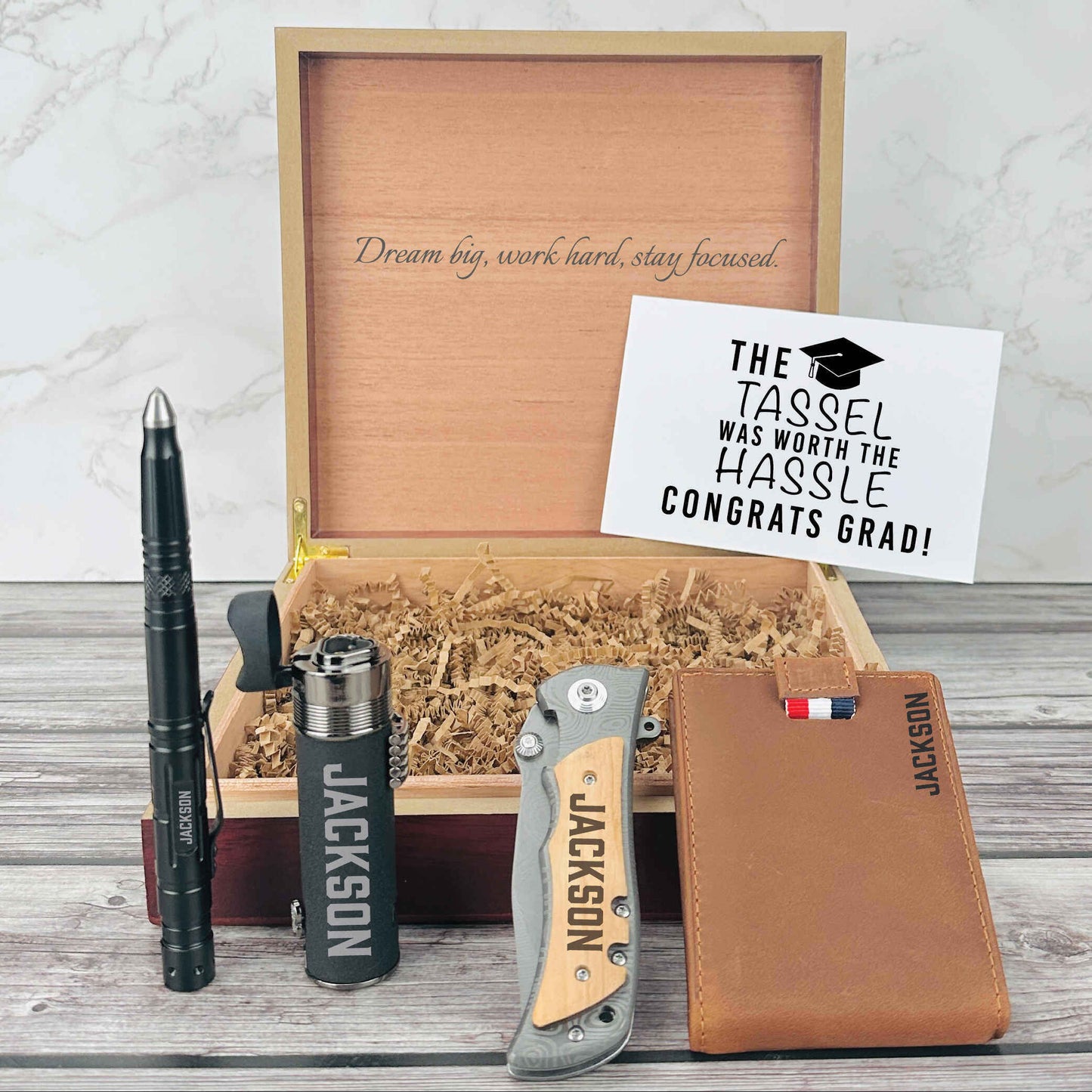 Wooden box with brass corners and engraved motivational text, containing personalized pen, flask, knife, and leather wallet with sample name engraving, displayed with graduation congratulations card.