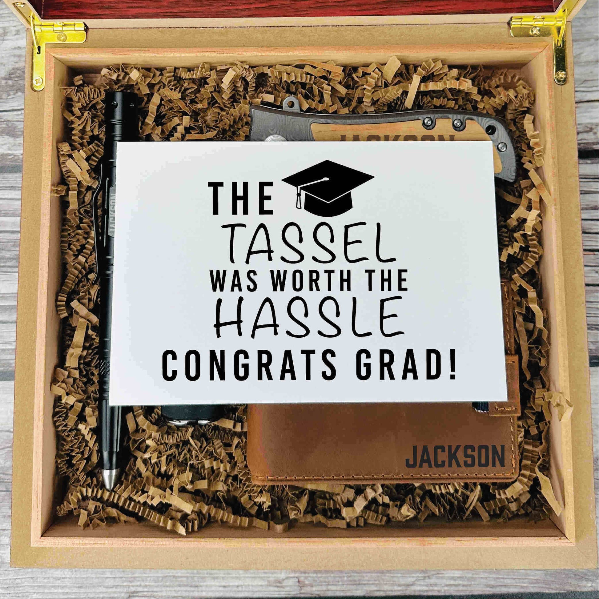 Wooden gift box with shredded kraft paper containing an engraved leather notebook with "JACKSON" and a black pen, displayed with a sample graduation card reading "The Tassel Was Worth the Hassle Congrats Grad!