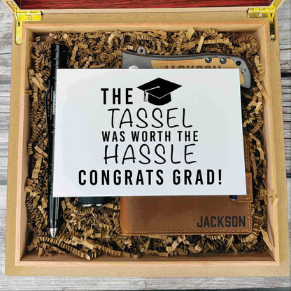 Wooden gift box with shredded kraft paper containing an engraved leather notebook with "JACKSON" and a black pen, displayed with a sample graduation card reading "The Tassel Was Worth the Hassle Congrats Grad!