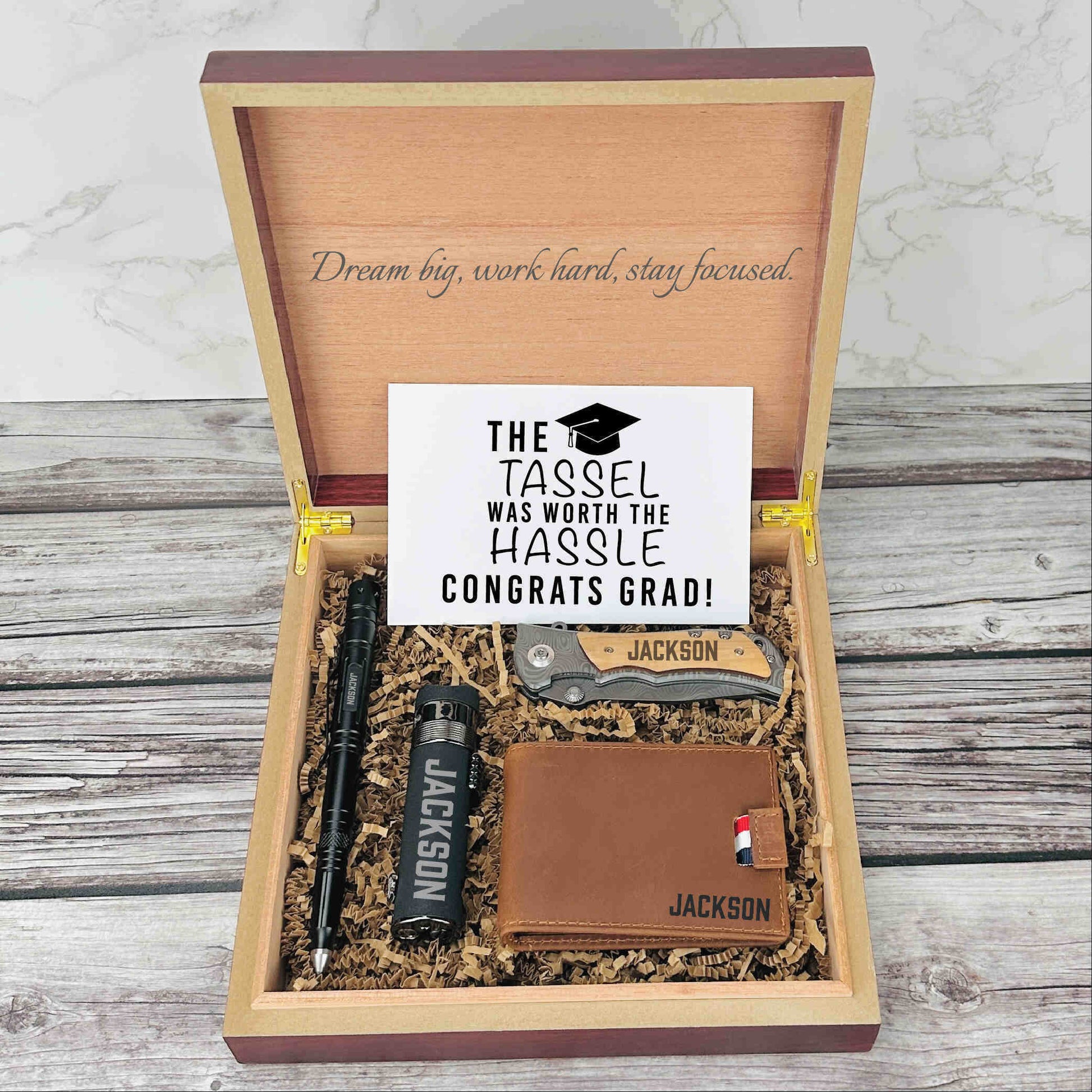 Wooden gift box with engraved motivational message, containing personalized knife, flask, wallet with sample name "Jackson," and congratulations card with graduation tassel design.