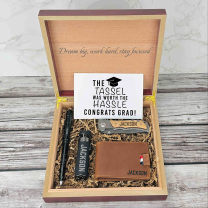 Wooden gift box with engraved motivational message, containing personalized knife, flask, wallet with sample name "Jackson," and congratulations card with graduation tassel design.
