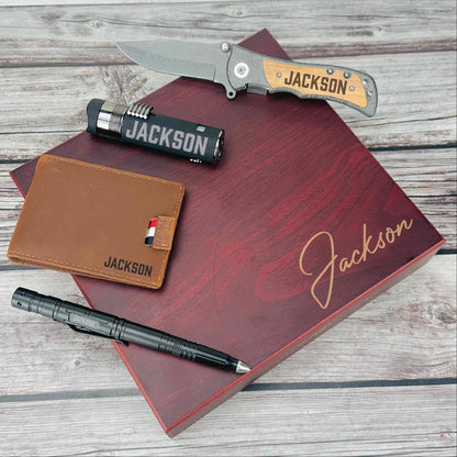Burgundy gift box with sample name "Jackson" engraved on lid, containing personalized folding knife with wooden handle, leather wallet, flashlight, and pen, displayed on wooden surface.