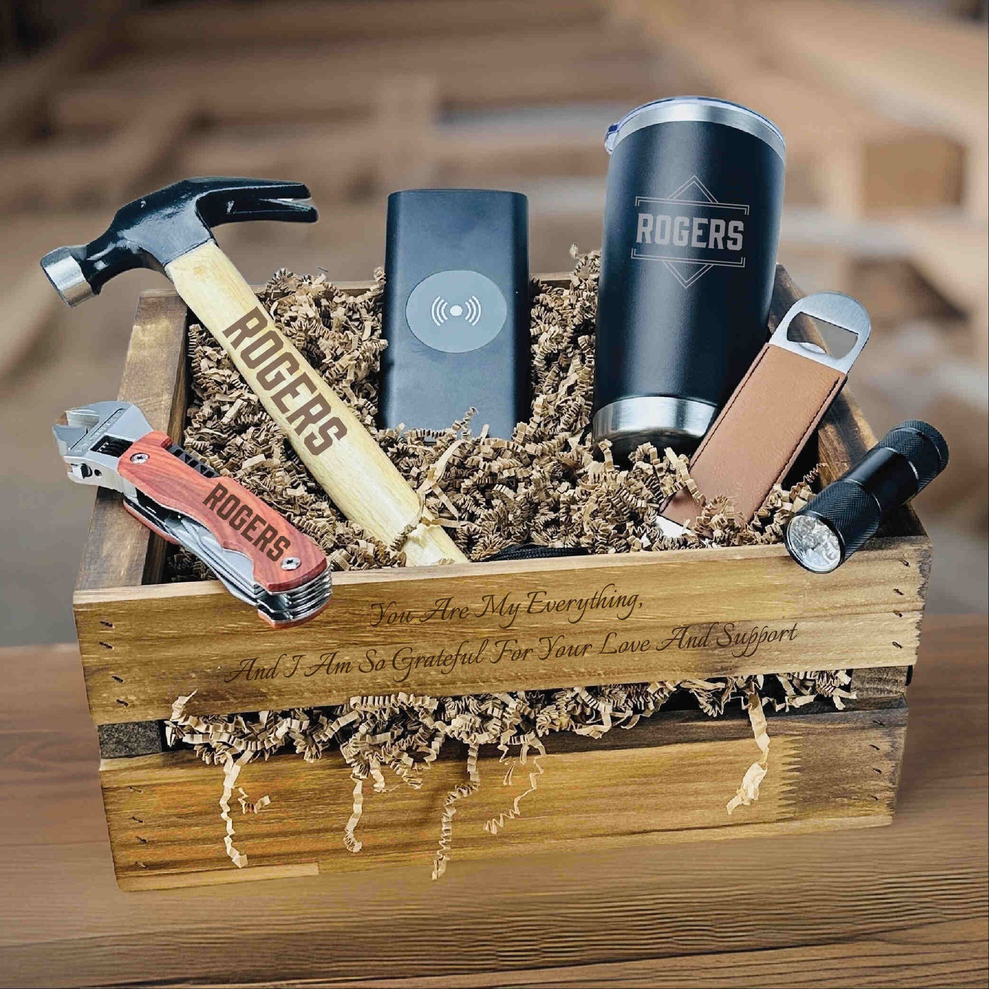 Wooden crate gift set with engraved items including hammer, pocket knife, flask, insulated tumbler, bottle opener, flashlight, and multi-tool, with sample personalization reading "You Are My Everything" on the box.