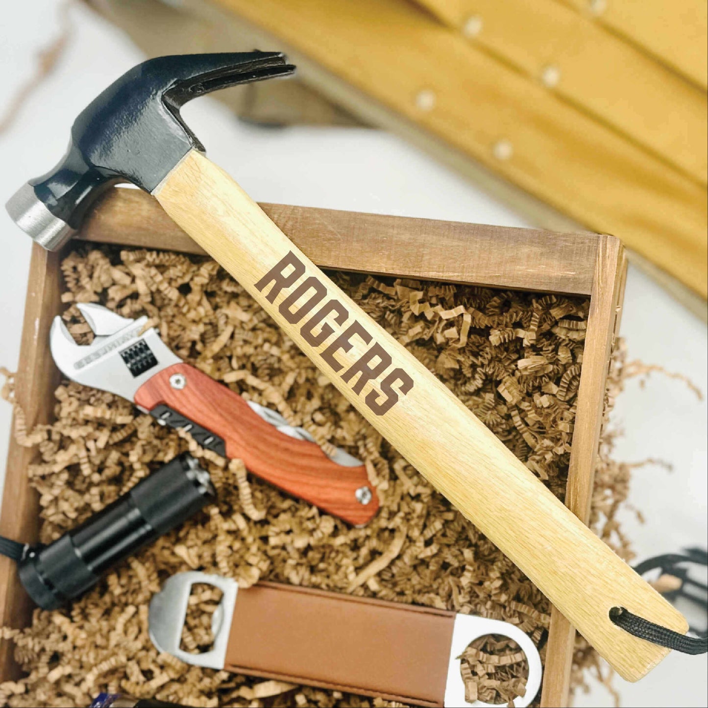 Engraved hammer with "ROGERS" sample text resting on wooden gift box filled with tools, wrench, and packing material.