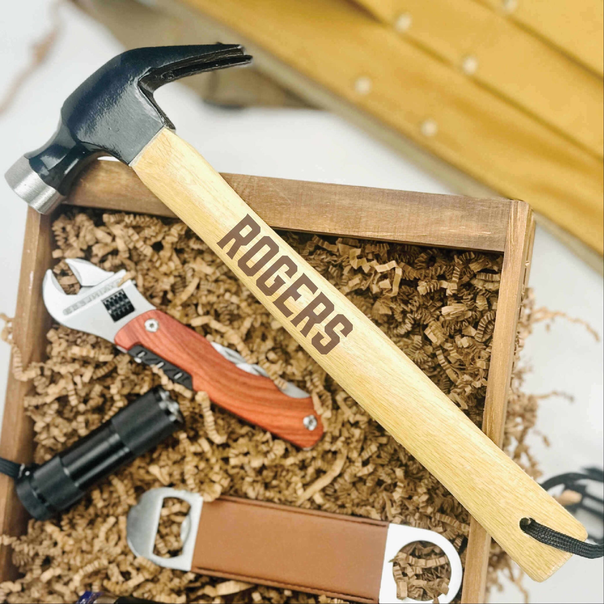Engraved hammer with "ROGERS" sample text resting on wooden gift box filled with tools, wrench, and packing material.